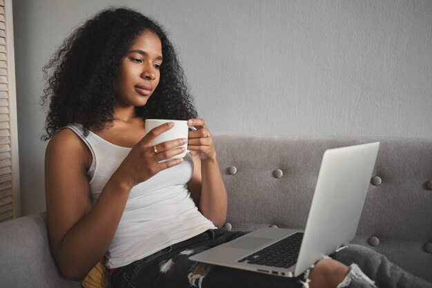 Tiro lateral de la atractiva joven mujer de piel oscura de moda en jeans rotos relajándose en el sofá con una computadora portátil en su regazo, tomando café y viendo series de televisión favoritas en línea