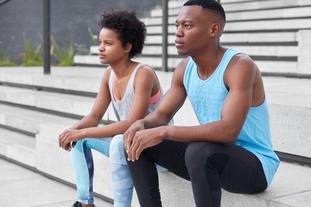 Tiro lateral de un corredor masculino de piel oscura vestido con ropa deportiva, se sienta cerca de su novia, posa en las escaleras, se enfoca en la distancia, toma un descanso después de los ejercicios físicos, tiene una forma de cuerpo deportivo. Deporte