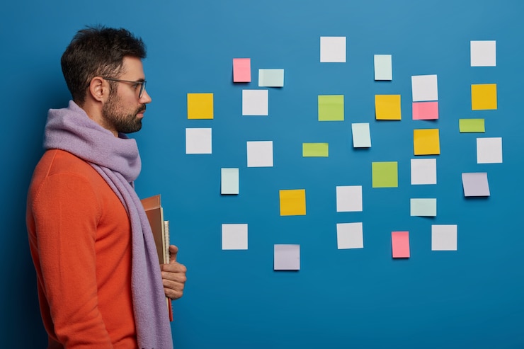 Foto gratuito tiro lateral de un estudiante barbudo serio que usa bufanda, jersey, sostiene libros, mira atentamente la pared azul con coloridas notas adhesivas piensa en las tareas del proyecto.