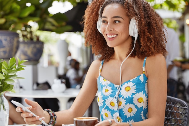 Tiro lateral de hermosa mujer afroamericana bastante sonriente sostiene un teléfono móvil moderno, escucha transmisiones de radio en línea, usa auriculares grandes blancos