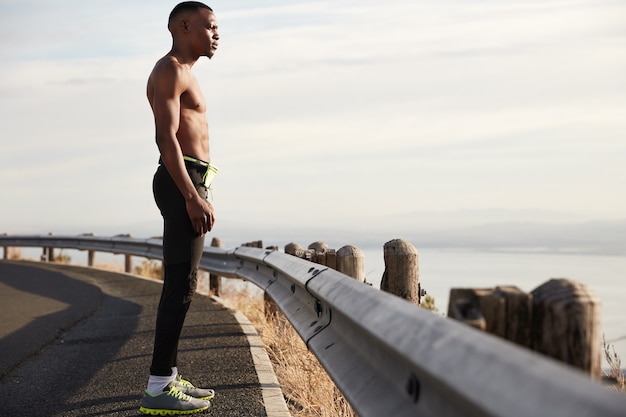 Tiro lateral de un hombre deportivo con piel oscura, tiene entrenamiento físico fuera de la ciudad, usa ropa deportiva, tiene una mirada seria y segura en la distancia, se para en el asfalto, se siente saludable. Personas y ejercicio