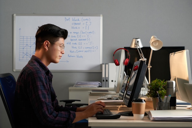 Tiro lateral del joven asiático en gafas trabajando en la computadora en la oficina