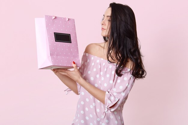 Foto gratuita tiro lateral de mujer joven seria morena mira la bolsa de regalo, usa un vestido de verano de moda, disfruta de recibir el presente, posa en rosa hembra hace compras, se encuentra en interiores