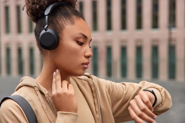 Tiro lateral de mujer rizada deportiva atenta comprueba el pulso después del entrenamiento concentrado en smartwatch vestido con poses de ropa deportiva al aire libre