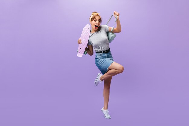 Tiro de longitud completa de niña en falda de mezclilla y camisa posando con longboard. Mujer joven tímida en traje gris y zapatillas blancas modernas mirando a la cámara.