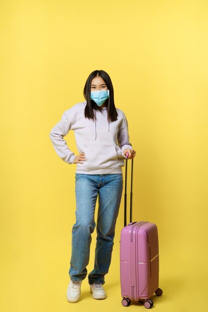 Tiro de longitud completa sonriente turista coreana en mascarilla médica, posando con maleta, ir de vacaciones, de pie sobre fondo amarillo.