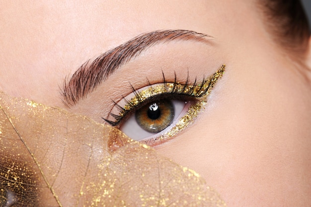 Tiro de macro de ojo femenino de belleza con maquillaje delineador de ojos dorado cubierto de hoja amarilla artificial