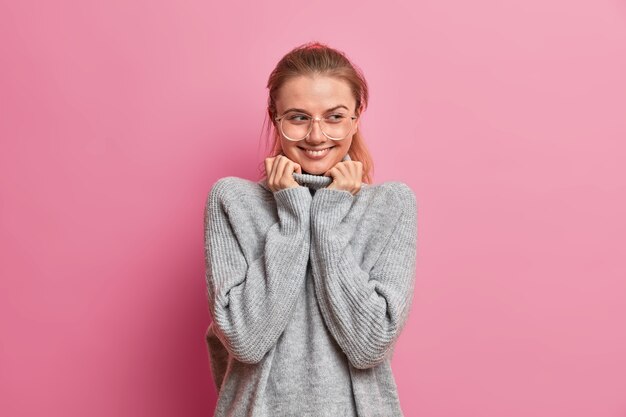 Tiro de media longitud de encantadora mujer adulta hermosa en suéter gris de gran tamaño sonríe tiernamente, recuerda algo agradable, usa gafas ópticas