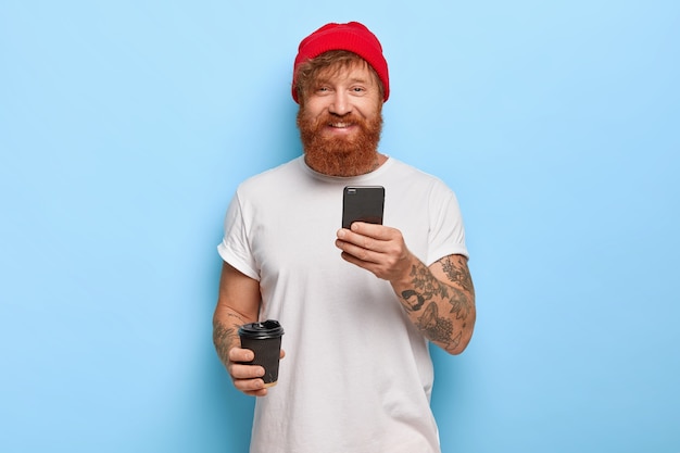 Tiro de media longitud de hombre alegre pelirrojo barbudo viste sombrero elegante, camiseta blanca casual, sostiene teléfono móvil, café para llevar, está de buen humor, escribe mensajes