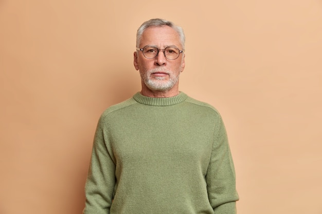 Tiro de media longitud de un hombre barbudo serio que parece impasible al frente con una expresión estricta, usa anteojos y el saltador tiene cabello gris y confía en algo aislado sobre la pared beige del estudio.