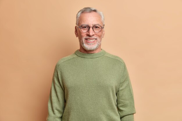 Tiro de media longitud de hombre mayor alegre sonríe felizmente con dientes blancos lleva gafas ópticas y un suéter aislado sobre una pared marrón