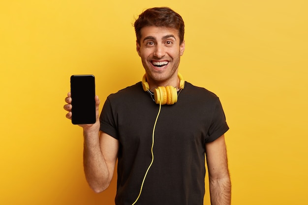 Tiro de media longitud de hombre optimista sostiene teléfono inteligente con pantalla simulada