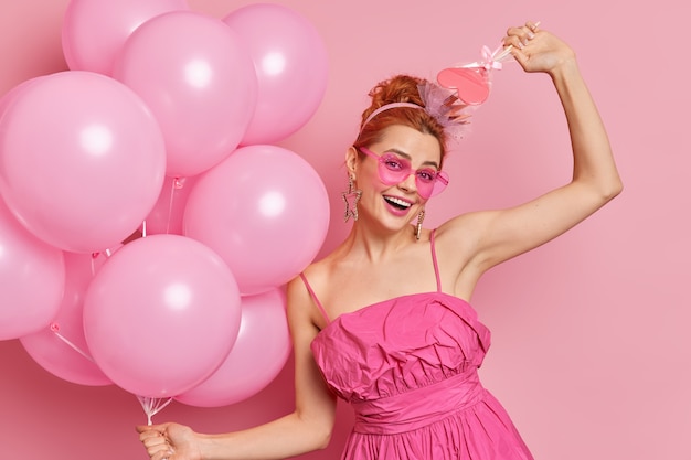 Tiro de media longitud de mujer europea alegre tiene bailes de humor alegre despreocupados con globos y dulces poses contra el fondo rosado en la fiesta.