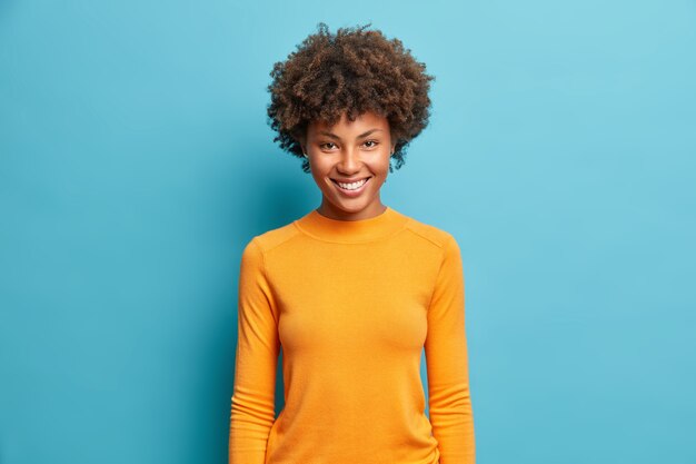 Tiro de media longitud de mujer joven bastante alegre con agradable sonrisa radiante expresión complacida vestida con un jersey naranja casual aislado en la pared azul