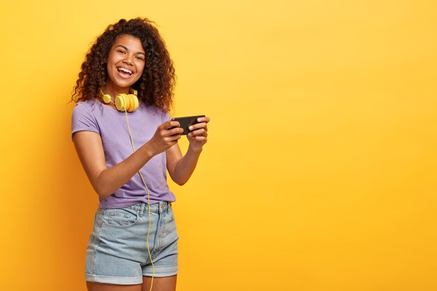 Tiro de media longitud de mujer joven positiva con peinado afro posando contra la pared amarilla