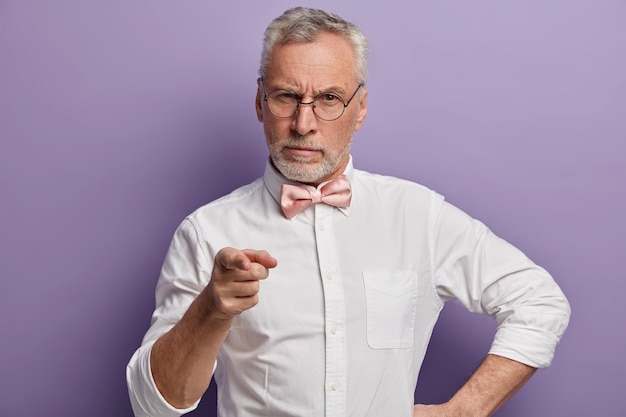 Tiro de media longitud de serio pensionista seguro de sí mismo con cabello gris, señala con el dedo índice a la cámara, usa gafas, elegante camisa blanca