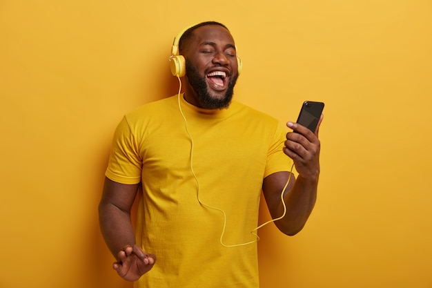 Tiro de medio cuerpo de chico negro escucha música para relajarse, sostiene un teléfono inteligente moderno y usa auriculares en las orejas, disfruta de una buena pista, posa sobre un fondo amarillo.