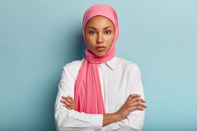 Tiro de medio cuerpo de una mujer seria y segura de sí misma que usa el tradicional hijab rosa, mantiene los brazos cruzados, se viste con una camisa blanca, se para contra la pared azul, escucha al interlocutor con interés. Religión