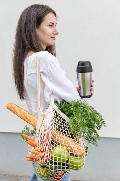 Tiro medio mujer mirando a otro lado y sosteniendo una bolsa reutilizable con comestibles y termo afuera