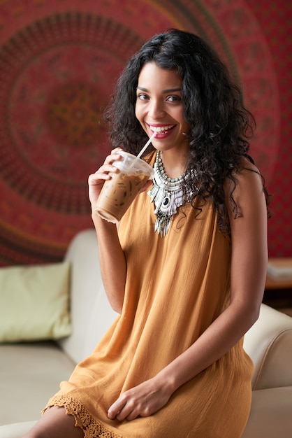 Tiro medio de mujer morena disfrutando de su café helado sentado en el sofá