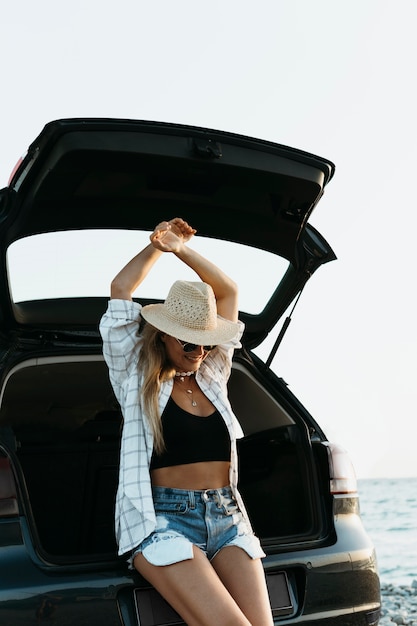 Foto gratuita tiro medio mujer rubia con la mano de pie en el maletero del coche con botella de jugo