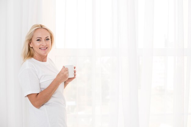 Tiro medio mujer sonriente con taza cerca de la ventana