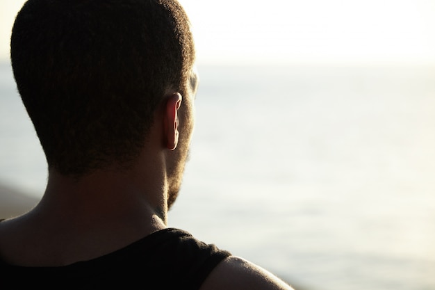 Foto gratuita tiro posterior del apuesto joven atleta de piel oscura que disfruta de los días soleados de verano en el océano, contemplando el amanecer