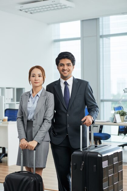 Tiro de rodilla de hombre y mujer en trajes posando en la oficina con maletas antes de un viaje de negocios