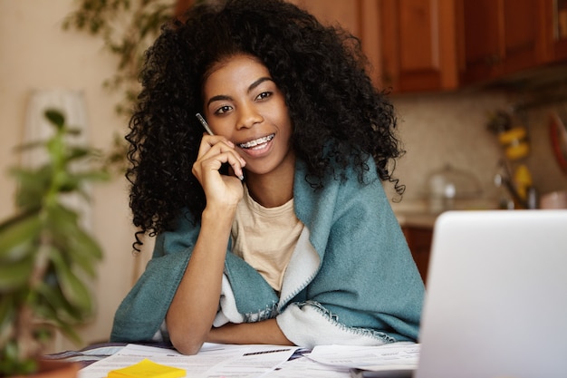 Tiro sincero de atractiva joven africana con pelo rizado vistiendo abrigo hablando por teléfono móvil