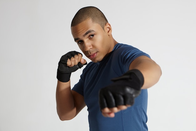 Tiro sincero de boxeador masculino de piel oscura joven serio musculoso con vendaje de boxeo y traje de entrenamiento practicando artes marciales en interiores. Culturista africano guapo fuerte trabajando en el gimnasio