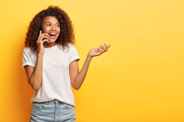 Tiro sincero de mujer joven despreocupada con pelo rizado posando con su teléfono