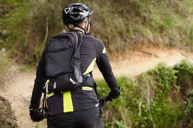 Tiro trasero del motorista en ropa de ciclismo negra y amarilla, casco y mochila montando bicicleta eléctrica de montaña en el camino mientras entrena al aire libre el fin de semana. Personas, estilo de vida saludable y concepto deportivo