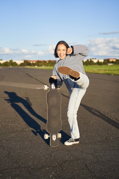 Foto gratuita tiro vertical de una chica asiática que se siente emocionada patinando en longboard saltando y posando con patineta