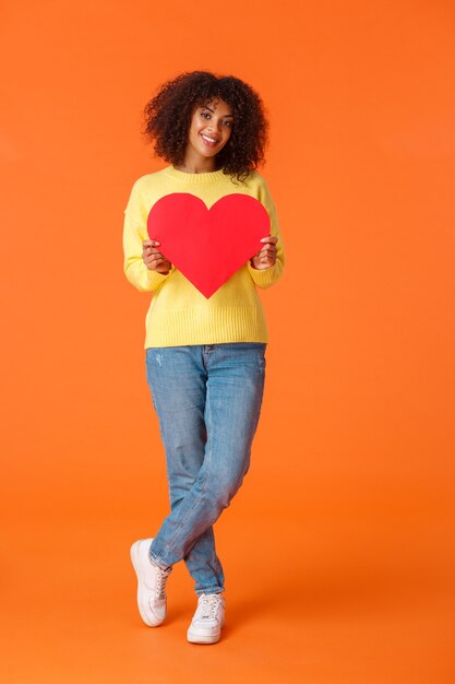 Tiro vertical de cuerpo entero hermosa, romántica y linda joven afroamericana con estilo y sosteniendo una gran tarjeta de corazón rojo para expresar amor, feliz día de San Valentín, confesar simpatía.