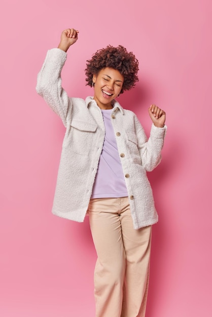 Tiro vertical de despreocupada mujer joven feliz con pelo rizado vestida con chaqueta de piel y pantalones baila con los brazos levantados mantiene los ojos cerrados aislados sobre la pared rosa