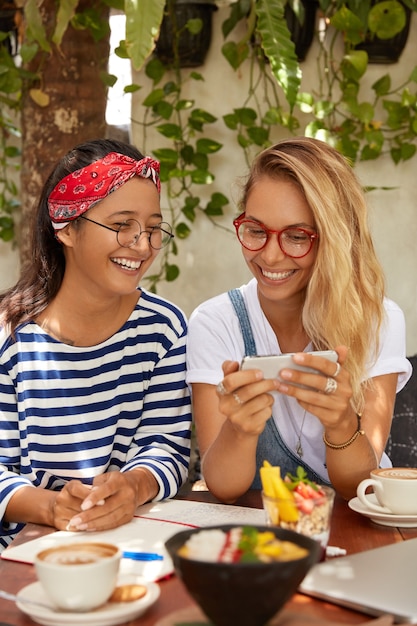 Tiro vertical de felices mujeres interraciales se ríen de buenos chistes, miran videos divertidos en teléfonos inteligentes