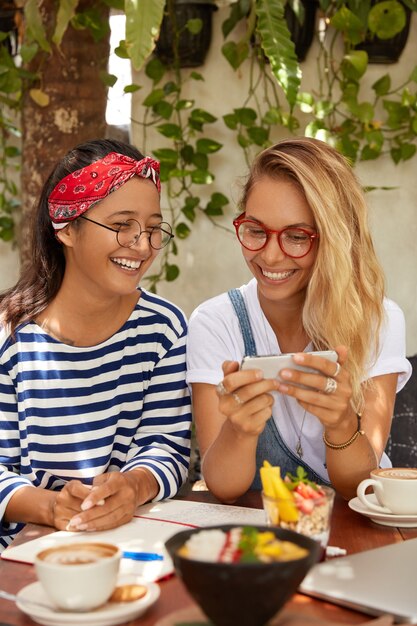 Tiro vertical de felices mujeres interraciales se ríen de buenos chistes, miran videos divertidos en teléfonos inteligentes