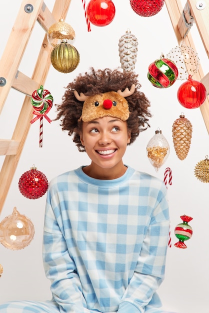 Tiro vertical de feliz soñadora mujer afroamericana tiene sueños de expresión pensativa sobre el milagro durante las vacaciones de invierno disfruta de un ambiente acogedor vestido con pijama contento de estar en casa. Víspera de año nuevo