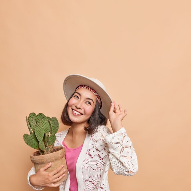 Tiro vertical de feliz soñadora mujer asiática con cabello oscuro compra cactus en maceta para el jardín de su casa tiene una expresión alegre usa fedora jersey blanco de punto posa contra el espacio en blanco de la pared beige