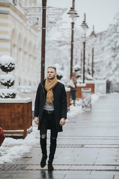Tiro vertical de un hombre sexy de moda caucásico con una bufanda marrón en el fondo de la calle nevada