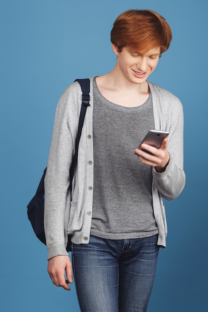 Tiro vertical de joven apuesto estudiante de jengibre en ropa gris y mochila sonriendo, charlando con un amigo por teléfono inteligente en camino a la universidad Estilo de vida moderno