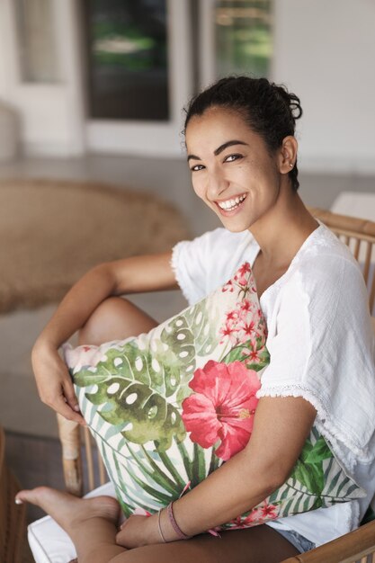 Tiro vertical linda mujer morena hispana en camiseta blanca, sentada con las piernas cruzadas en una cómoda silla de ratán