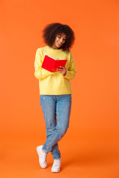 Tiro vertical de longitud completa soñadora linda estudiante afroamericana con corte de pelo afro, ropa casual, yendo a la universidad, tomando notas, de pie sobre una pared naranja y escribiendo en un cuaderno sonriendo.