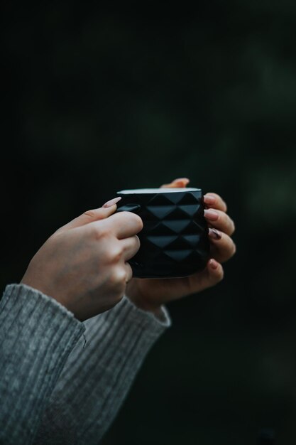 Tiro vertical de manos femeninas en un suéter acogedor sosteniendo una taza de café negro