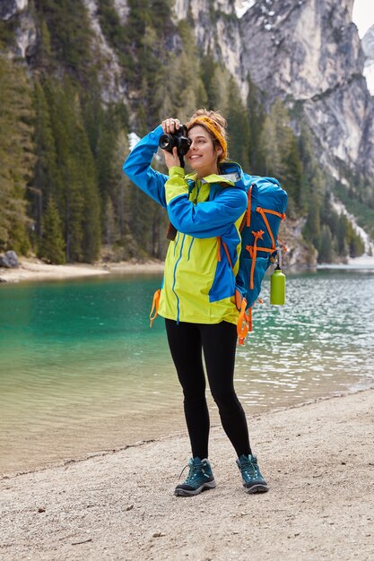 Tiro vertical de mujer alegre hace fotos profesionales del paisaje de la naturaleza, lleva chaqueta