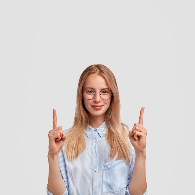 Tiro vertical de mujer europea joven satisfecha con expresión complacida, apunta hacia arriba con los dedos índices, vestida con camisa de moda, muestra algo arriba, aislado sobre una pared blanca.