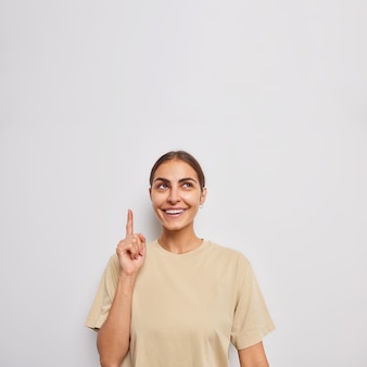 El tiro vertical de la mujer europea positiva apunta arriba con el dedo índice muestra publicidad u oferta promocional vestida con una camiseta casual aislada sobre una pared blanca elige algo en la tienda