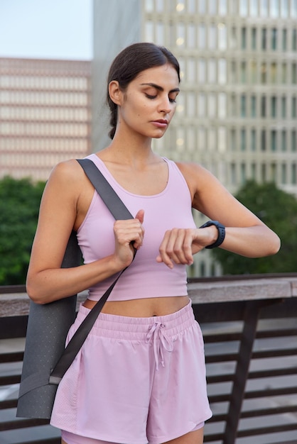 Tiro vertical de mujer joven delgada deportiva en ropa deportiva comprueba el tiempo en el reloj inteligente espera a que el entrenador comience el entrenamiento físico o las posturas de práctica de yoga en el puente