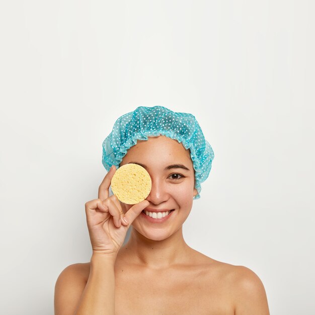 Tiro vertical de mujer joven feliz mantiene la esponja en el ojo, tiene tratamientos de belleza, quita el maquillaje antes de tomar la ducha