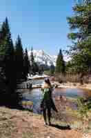 Foto gratuita tiro vertical de una mujer con mochila de pie cerca del agua y los árboles con una montaña en segundo plano.